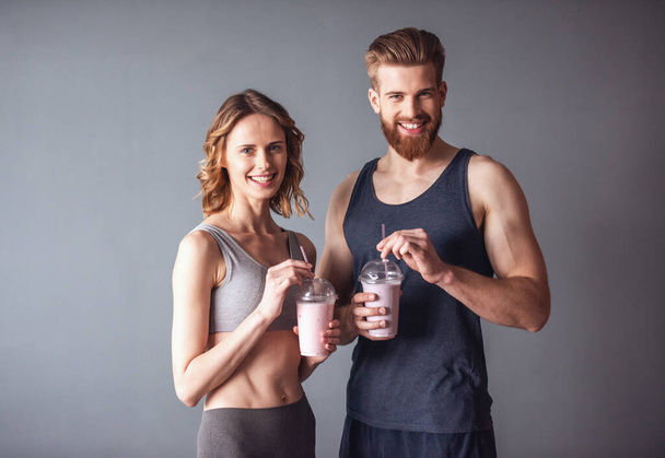 Beautiful young sports people are holding nutritious cocktails, looking at camera and smiling, on gray background - Photo, Image