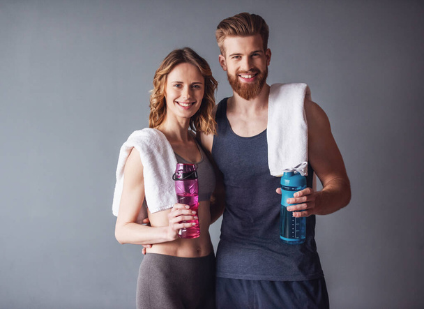 Beautiful young sports people are holding bottles of water, looking at camera and smiling, on gray background - Photo, Image