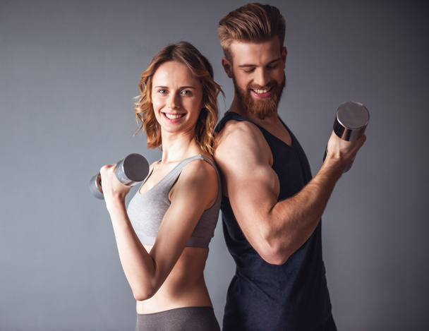 Beautiful young sports people are working out with dumbbells and smiling, on gray background - Photo, Image