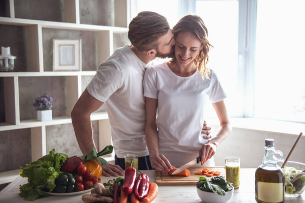 Όμορφο νεαρό ζευγάρι να μιλάμε και να χαμογελά μαγειρεύοντας υγιεινά τρόφιμα στην κουζίνα στο σπίτι. Ο άνθρωπος φιλιά την κοπέλα του στο μάγουλο - Φωτογραφία, εικόνα