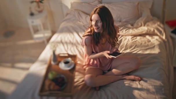 Romantikus nő, aki okostelefont gépel a reggeli napfényben. Nyugodt lány kényelmes ágyban pihen sms-ező mobiltelefon. Boldog gyönyörű modell élvezze a hétvégét a szállodai szobában. Pozitív női ellenőrző eszköz pizsamában - Felvétel, videó
