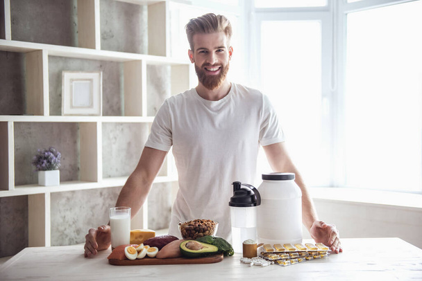 Handsome young bearded sportsman is looking at camera and smiling while preparing sport nutrition in kitchen at home - Photo, Image