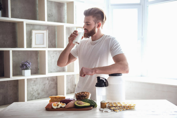 Красивый молодой бородатый спортсмен пьет во время подготовки спортивного питания на кухне дома
 - Фото, изображение