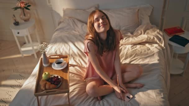 Dromerig meisje die 's morgens in bed ligt. Happy teenager holding smartphone genieten weekend ontbijt. Een serene glimlachende vrouw die haar ogen sluit droomt in een knusse hotelkamer. Mooi romantisch vrouwtje thuis - Video