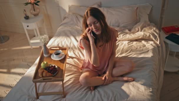 Pozitív női beszélő mobiltelefon az ágyban. Nyugodt lány pihen napos reggel pizsamában élvezi a reggelit. Mosolyogó nyugodt nő beszél befejező hívás otthon. Boldog gondtalan tinédzser tervezési hétvégét - Felvétel, videó