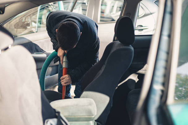 One young handsome caucasian brunette man vacuuming the front passenger seat inside the salon in his car at the city car wash on a clear spring sunny day, top view, close-up. - Photo, Image