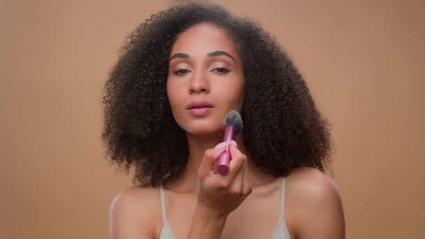 Afroamerykanka stosując proszek makijaż naturalne piękno czyste miękkie nawilżona nawilżona pielęgnacja skóry leczenie dziewczyna zrobić rumieniec na twarz brąz konturowanie za pomocą makijażu pędzel kosmetyczne beżowe tło - Materiał filmowy, wideo