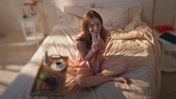 Happy teenager holding smartphone resting in sunny bedroom. Dreamy model enjoy morning checking browsing mobile phone in cozy bed. Romantic smiling girl rest at home pajamas. Carefree leisure concept - Footage, Video