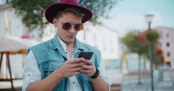 Uśmiechnięty młodzieniec noszący kapelusz i okulary przeciwsłoneczne SMS-y na telefonie komórkowym - Materiał filmowy, wideo