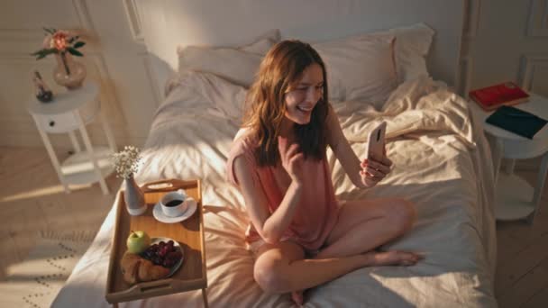 Chill girl rozmawia o wideokonferencji w słonecznej sypialni. Szczęśliwy romantyczny nastolatek machając kończąc połączenie online trzymając smartfona. Beztroska piękna kobieta odpoczywa rano w domu. Weekendowy styl życia śniadanie - Materiał filmowy, wideo