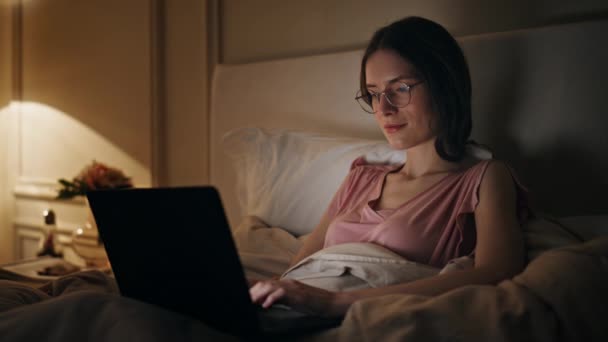Abendmädchen tippt Laptop im Bett. Entspannte lächelnde Frau, die im Internet surft und sich im gemütlichen Schlafzimmer ausruht. Konzentrierte Freiberufler überarbeiten das Online-Shopping zu Hause. Abendunterhaltung am Wochenende. - Filmmaterial, Video