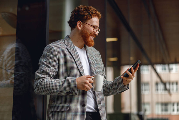 Χαμογελώντας επιχειρηματίας κρατώντας τηλέφωνο, ενώ στέκεται στο γραφείο κατά τη διάρκεια του διαλείμματος. Υψηλής ποιότητας φωτογραφία - Φωτογραφία, εικόνα