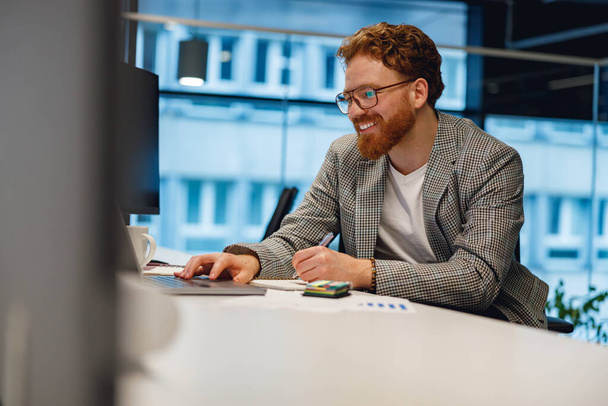 Улыбающийся мужчина-менеджер, работающий над ноутбуком, сидя за столом на фоне офиса - Фото, изображение