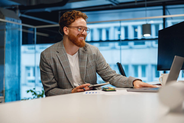 Χαμογελώντας αρσενικό διαχειριστή που εργάζονται σε φορητό υπολογιστή, ενώ κάθεται στο γραφείο στο φόντο του γραφείου  - Φωτογραφία, εικόνα