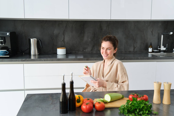 Retrato de la mujer anotando la lista de comestibles, tomando notas en la receta, sentado en la cocina cerca de las verduras, preparando el menú de la cena. - Foto, Imagen