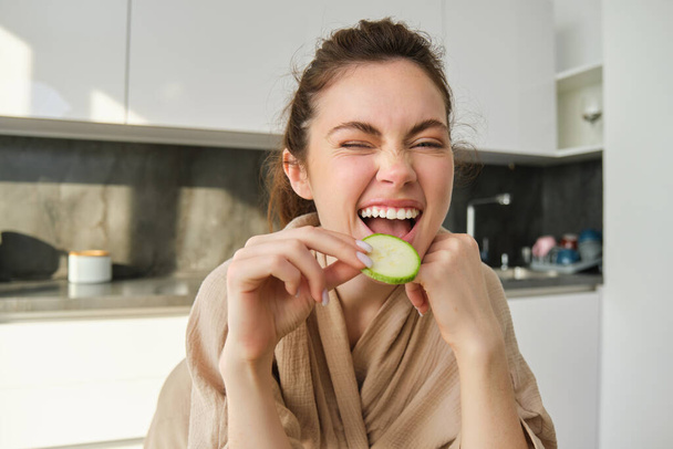 Retrato de una joven feliz y sonriente en la cocina, cocinando, picando calabacín, sosteniendo verduras y luciendo feliz, preparando comida vegana en casa. - Foto, imagen
