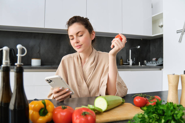 Beeld van jonge mooie vrouw, met tomaat, zittend in de keuken met smartphone, snijplank en groenten op de toonbank, koken eten, boodschappen voor haar recept, met behulp van mobiele telefoon app. - Foto, afbeelding