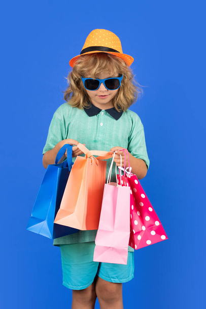 Slevy a prodeje. Šťastné dítě s nákupními taškami. Dítě s nákupními taškami pózující na modrém studiu izolované pozadí. Trendy a značky - Fotografie, Obrázek