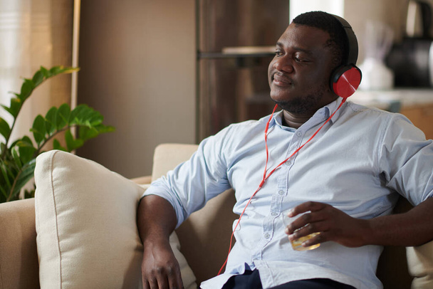 Retrato del hombre feliz bebiendo un vaso de whisky y escuchando música en auriculares - Foto, imagen