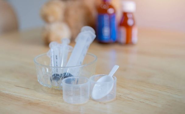 Focus selettivo di cucchiai di plastica medica e siringa per bambini sul tavolo di legno con offuscata una bottiglia di vitamine terra nera. Concetto di medicina tradizionale bambini. - Foto, immagini