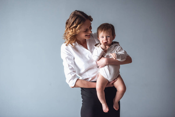 Ωραία μαμά σε κοστούμι παίζει με το χαριτωμένο μωρό αγόρι και χαμογελώντας, σε γκρίζο φόντο - Φωτογραφία, εικόνα