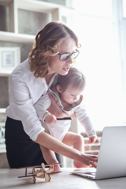 Hermosa mamá de negocios está utilizando un ordenador portátil mientras pasa tiempo con su lindo bebé en casa
 - Foto, Imagen