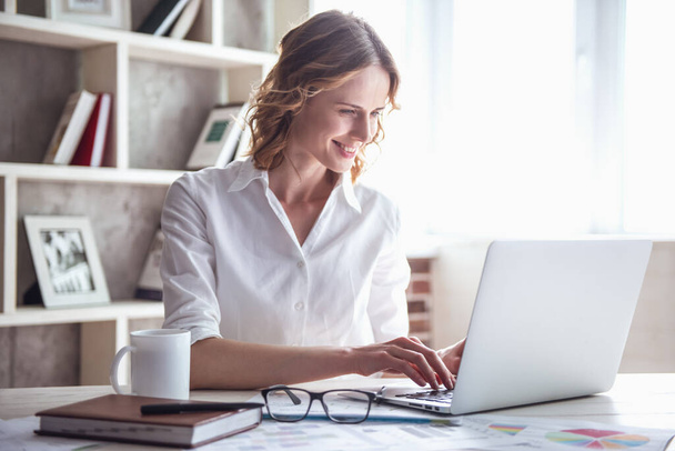 Bella donna d'affari sta usando un computer portatile e sorridente mentre lavora in ufficio
 - Foto, immagini