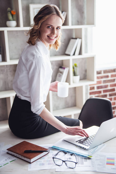 Beautiful business woman is drinking coffee, using a laptop and smiling while working in office - Photo, Image