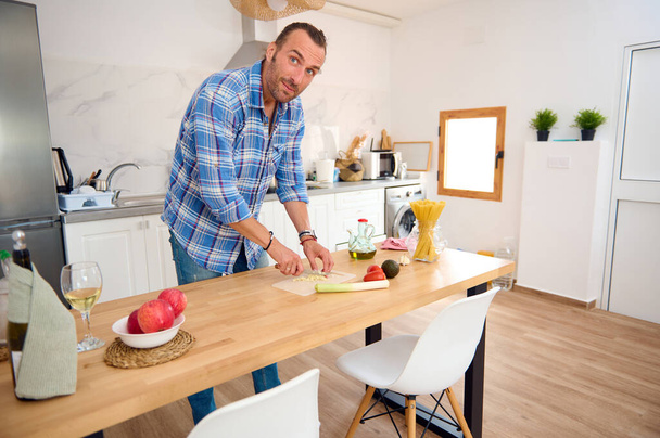 Молодой белый мужчина в повседневной одежде, улыбается, смотрит в камеру, режет чеснок, готовит соус для итальянской пасты на домашней кухне. Здоровый образ жизни, питание и диета - Фото, изображение
