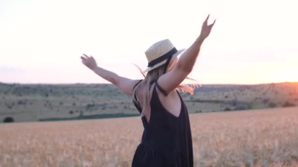 szczęśliwa kobieta w średnim wieku w czarnej sukience i kapeluszu biegnie w świetle zachodu słońca na polu pszenicy - Materiał filmowy, wideo