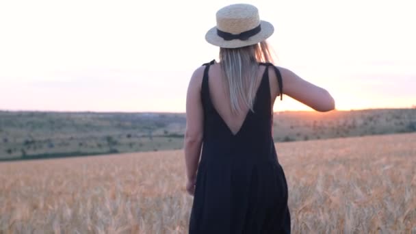 Ευτυχισμένη μεσήλικη γυναίκα με μαύρο φόρεμα και καπέλο τρέχει στο φως του ηλιοβασιλέματος στον τομέα του σιταριού - Πλάνα, βίντεο