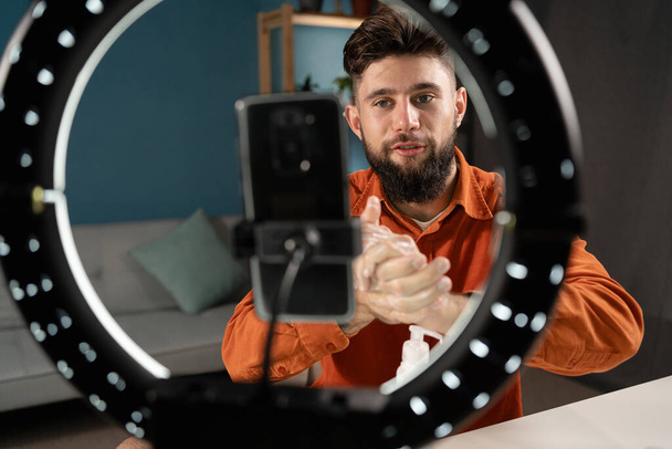 blogueiro de beleza masculino com vídeo de gravação de produto cosmético na sala de estar usando smartphone e lâmpada de anel. Espaço de cópia - Foto, Imagem