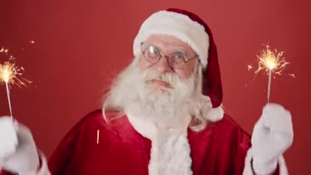 Mediano primer plano de Santa Claus alegre en gafas, con barba y bigote sosteniendo chispas de fuegos artificiales ardientes en ambas manos y bailando enérgicamente, sobre fondo rojo. Plantilla, espacio de copia - Imágenes, Vídeo