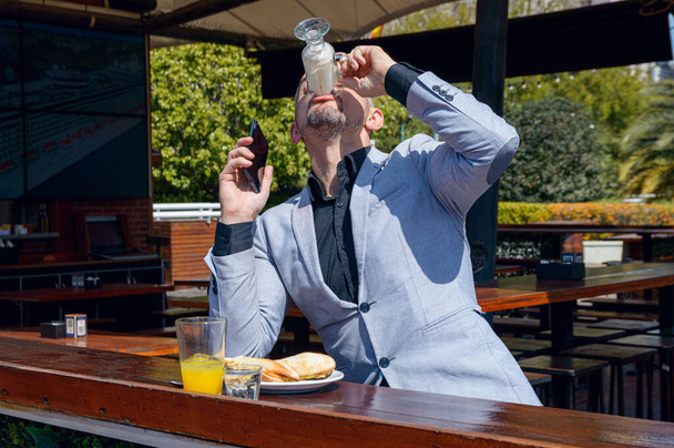 Hombre de negocios adulto caucásico, vestido con ropa elegante, sentado con el teléfono en la mano, terminando su capuchino, restaurante al aire libre, desayunando por la mañana antes de empezar a trabajar. - Foto, imagen