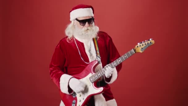 Tiro médio de homem idoso barbudo em traje de Papai Noel, com óculos de sol e corrente de prata, energeticamente tocando guitarra elétrica, em fundo vermelho liso. Modelo, espaço de cópia - Filmagem, Vídeo