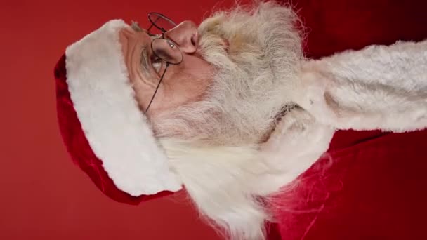 Lähikuva pystysuora kuva kasvoista Santa punainen hattu ja lasit, paksu parta ja viikset, katsot kameraa ja laittaa sormi huulille, kehottaen salailua tai hiljaisuutta. Malli, kopiotila - Materiaali, video