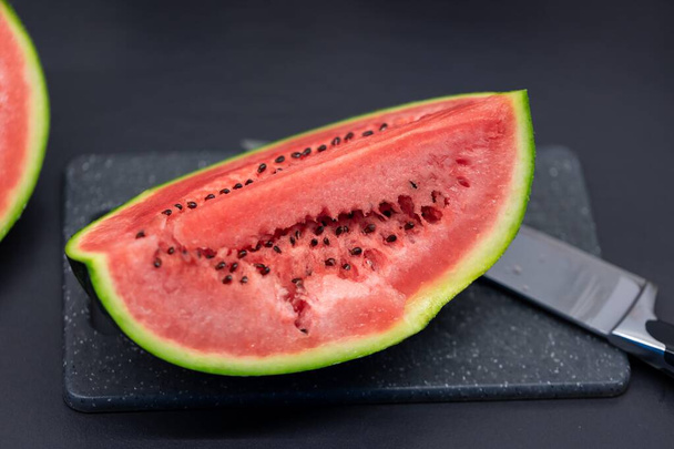 Das Porträt eines Viertelschnitts einer grünen Wassermelone liegt auf einem Schneidebrett aus Kunststoff neben einem Messer. Das Stück oder die Scheibe der Frucht ist fertig zum Verzehr und man sieht schwarze Samen im roten Fruchtfleisch. - Foto, Bild