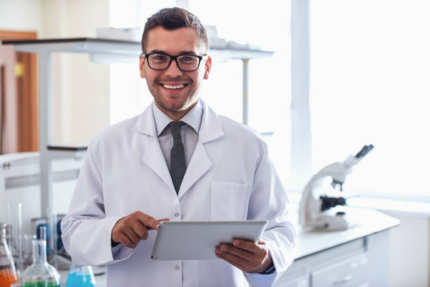 schöner Arzt im weißen Kittel benutzt ein digitales Tablet, blickt in die Kamera und lächelt im Labor - Foto, Bild