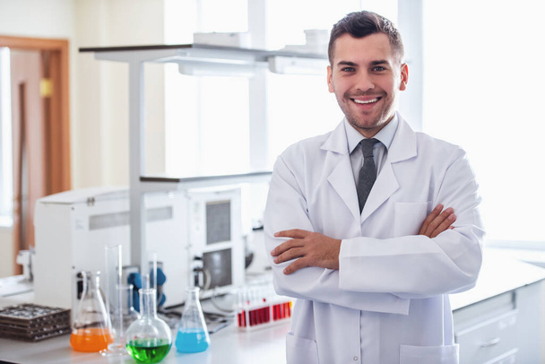 Handsome medical doctor in white coat is looking at camera and smiling while standing in laboratory - Photo, Image