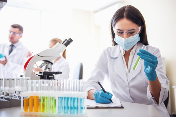 Όμορφη γιατρός γάντια και μάσκα κάνει σημειώσεις ενώ εργάζεστε με σωλήνες δοκιμής στο εργαστήριο - Φωτογραφία, εικόνα