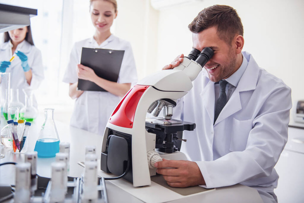 schöner Arzt benutzt ein Mikroskop und lächelt, während er im Labor arbeitet - Foto, Bild
