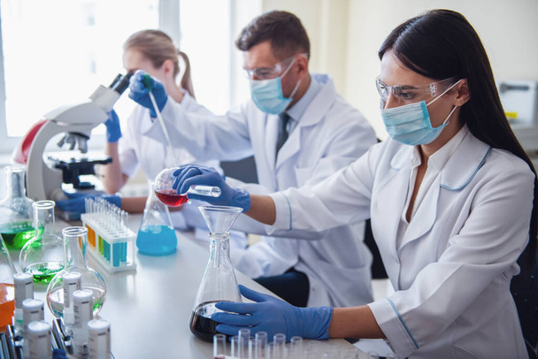 Schöne Ärzte in Handschuhen, Masken und Brillen arbeiten im Labor mit Substanzen in Reagenzgläsern und Mikroskop - Foto, Bild