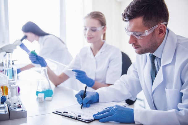 Belos médicos em luvas e óculos estão trabalhando com substâncias em tubos de ensaio e microscópio no laboratório
 - Foto, Imagem