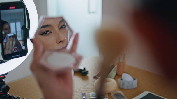 Influencer žena nanášení make-up vypadající zrcadlo v domě detailní. Beauty blogger uvedení nadace natáčení vizualizace rutinní výuka na mobilní telefon nastavení na ring light stand. Dáma přezkoumávající kosmetiku - Fotografie, Obrázek