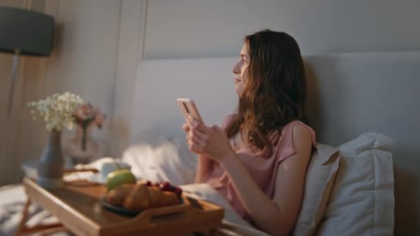 Zrelaksowana dziewczyna pisząca wiadomość w domowym łóżku. Atrakcyjna wymarzona kobieta, leżąca na telefonie w weekendowy poranek. Spokojny uśmiech kobiety korzystających śniadanie zakupy online. Codzienna rutyna za pomocą aplikacji - Materiał filmowy, wideo