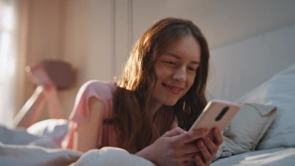Όμορφη κοπέλα που ψάχνει στα μέσα κοινωνικής δικτύωσης στο κοντινό κρεβάτι. Ευτυχισμένη νεαρή γυναίκα ξεκουράζεται σπίτι δακτυλογραφώντας κινητό τηλέφωνο το πρωί. Ξέγνοιαστη χαρούμενη γυναίκα που χρησιμοποιεί μηνύματα smartphone online. Χαρούμενη οθόνη μοντέλου. - Πλάνα, βίντεο