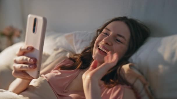 Menina fazendo videochamada na cama closeup. Sorrindo mulher feliz acenando smartphone falando em conferência online. Acorde alegre videoclipe feminino descansando à luz do sol da manhã. Conceito de relação à distância - Filmagem, Vídeo