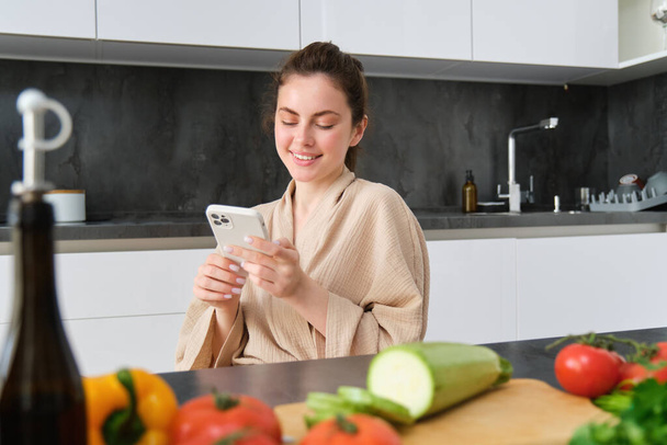スマートフォンでレシピを見ている女性, 野菜やチョッピングボードとキッチンに座って, 夕食の準備, 健康的なサラダ, 自宅で料理, バスローブを着用. - 写真・画像