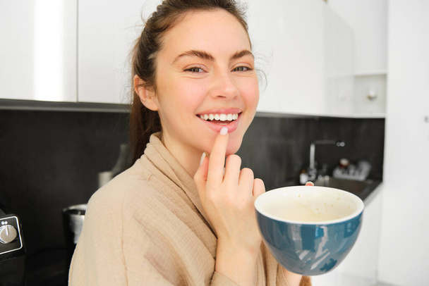 Primer plano retrato de chica atractiva bebiendo café, sosteniendo taza con cappuccino de la mañana y sonriendo, teniendo una taza de deliciosa bebida en la cocina, usando cómodo albornoz. - Foto, Imagen