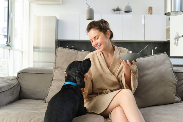 Conceito de casa e estilo de vida. Mulher bonita com cão, sentado no sofá, comendo cereais e abraçando seu filhote de cachorro, menina tomando café da manhã em roupão. - Foto, Imagem